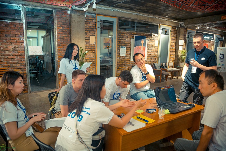 Participants at the Ecommerce and Fintech Hackathon 2019 in Bishkek, Kyrgyzstan, gather around a table with a laptop and documents.