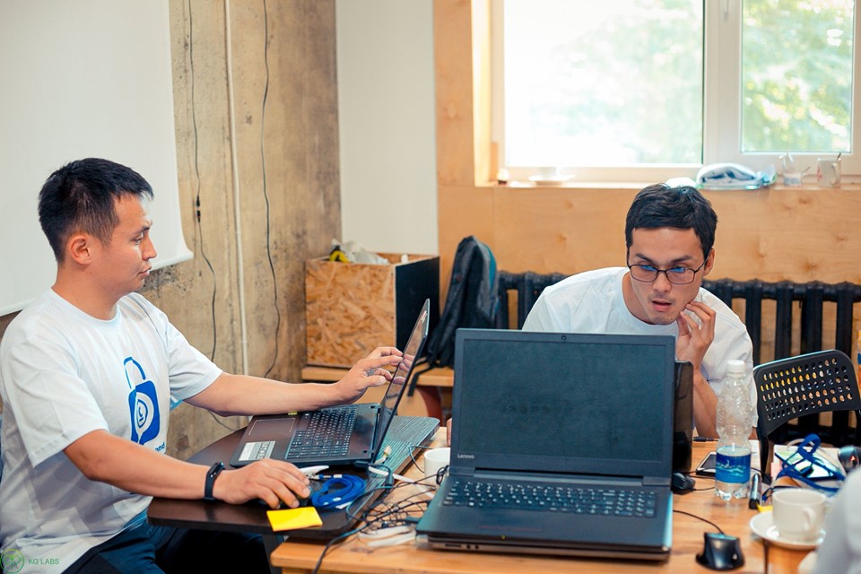 Two men working on laptops at a table in a workshop setting.