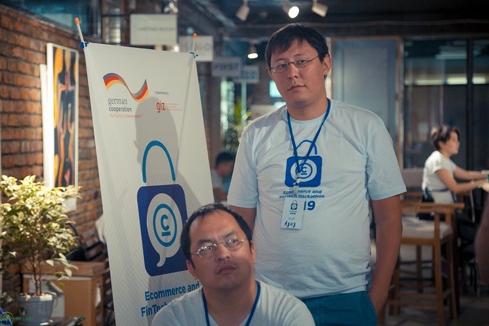 Two men pose in front of a banner for the Ecommerce and Fintech Hackathon 2019 in Bishkek, Kyrgyzstan.