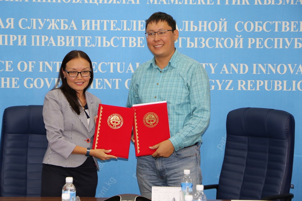 Two individuals holding red folders at a table in front of a blue background with text.
